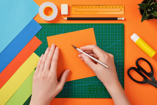 Hands working with colorful paper using a precision knife on a green cutting mat. Tools for paper crafts. Ruler, pencil, glue stick, scissors, tape, eraser