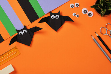 Origami paper bats with goggle eyes on a orange background. Surrounded by crafting supplies, paper...