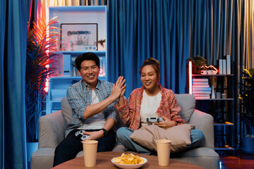Couple gamer making high five holding joysticks be winner together complete level sitting sofa playing video game with front snack and drinks at neon light color living room at modern home. Infobahn.