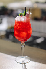 Red cocktail with ice served in wine glass. Garnished with mint. Straw and skewer. Refreshing summer drink. Blurred bar background. Cold beverage on counter. Bright sunlght vibrant alcohol.