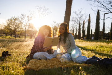 Two female students sitting on a blanket in a park, sharing snacks and drinks while engrossed in a book together during a beautiful sunset, creating a moment of friendship and relaxation