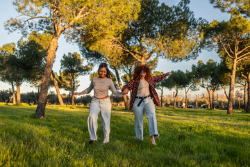 Fototapeta premium Two carefree young women are running through a grassy park holding hands and smiling, enjoying the warm sunlight filtering through the trees