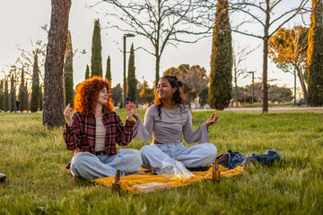 Two diverse young women are sitting in a lotus position on a blanket in a park, practicing meditation and enjoying each other's company during a relaxing sunset picnic
