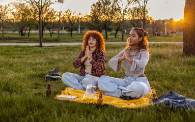 Two female friends are practicing yoga and meditating in a park at sunset, sitting on a blanket with snacks and drinks nearby, enjoying a peaceful moment after their workday