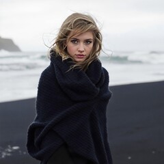 Portrait of a serious young woman wrapped in a large knit scarf, standing on a black sand beach with rough sea background.