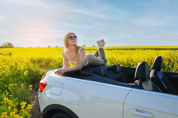 Young woman relaxing on a convertible car in the middle of a yellow rapeseed field at sunset.