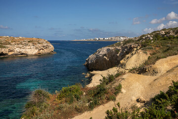 Scenic coastal landscape with wild vegetation on rocky slopes, turquoise sea, and distant town under a blue sky. Mediterranean nature for travel, summer, and nature themes.
