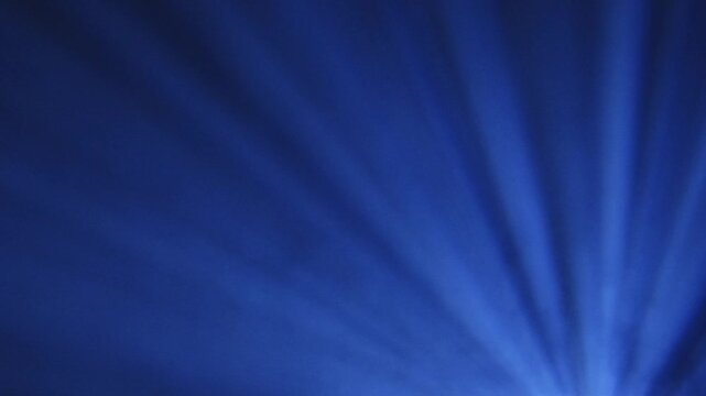 Bright blue spotlight projecting rays through haze or smoke, creating dramatic beams of light spreading outward in a dark environment with copy space.