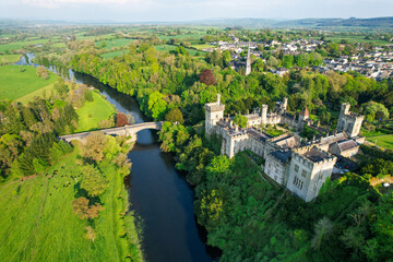  Aerial view of historic Lismore Castle and the surrounding townscape in County Waterford, Ireland, with lush spring greenery, the River Blackwater, and rolling countryside under soft evening light.