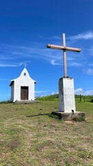 Saint Peter Chapel, a tiny whitewashed chapel built by fisherman on the top of a mound, and its front wooden cross. Blue sky, white clouds, green lawn. Fernando de Noronha island, Brazil. Sunny day.