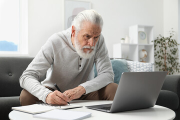 Fototapeta premium Senior man taking notes while learning online at table indoors. Self-study