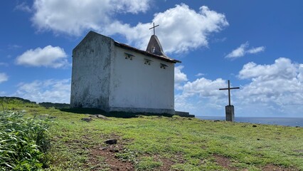 Saint Peter Chapel, a tiny whitewashed chapel built by fisherman on the top of a mound, and its front wooden cross. Blue sky, white clouds, green lawn. Fernando de Noronha island, Brazil. Sunny day.