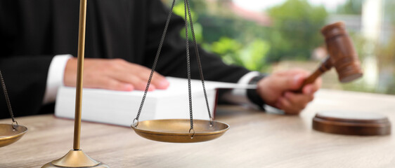 Closeup of judge with law book and gavel at table in office, focus on scales of justice. Banner...