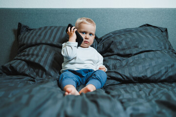 Obraz premium Boy 2 years old with a smartphone. Toddler boy sitting in bed and playing with a mobile phone. Calling mom. Cute boy holding a mobile phone in his hands and looking intently at the screen.