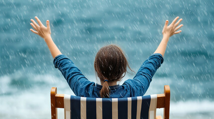 Rear view of a woman in a striped chair enjoying rainfall by the sea. Arms raised in joy, capturing freedom and nature's refreshing embrace.