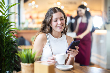 Young woman in cafe drinks coffee and using mobile phone, scrolls through social media feed, chats online, calls friend, talks on phone. Free Wi-Fi zone, Internet cafe.