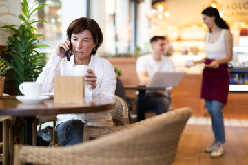 Elderly woman talking on mobile phone and drinking coffee in cafe