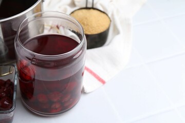 Tasty cherry compot in glass jar and ingredients on white tiled table, closeup. Space for text