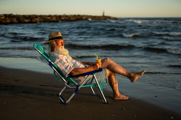 Thoughtful elderly man in striped shirt and straw hat sitting on beach chair by the sea, holding orange juice, peacefully watching ocean waves and enjoying a moment of calm during golden hour.