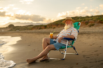 Elderly man with white beard and straw hat relaxing in a beach chair, holding orange juice, enjoying peaceful retirement and tranquil sunset atmosphere by the sea on a warm summer evening...