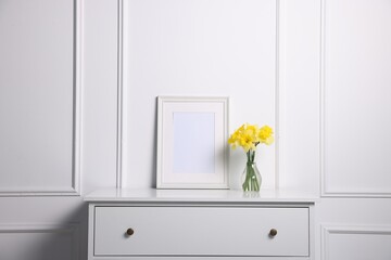 Beautiful daffodils in vase and picture frame on cabinet near white wall at home