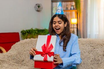 Indian young woman opens birthday gift while on home sofa and reacts with amazed expression and big...