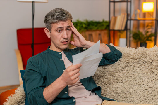 Young sad man at home opens envelope and sees credit bill, wipes forehead and frowns. Caucasian unhappy guy on sofa expresses visible concern, emotional reaction and disappointment about high payment
