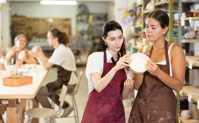Interested young girls wearing aprons viewing attentively handmade clay bowl in pottery workshop