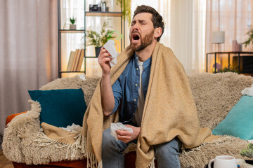 Young sick ill man at home sits on the sofa, wrapped in blanket, sneezing and quickly reaching for...