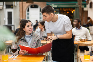Male waiter takes an order from a young woman on the veranda of a restaurant, he asks preferences...