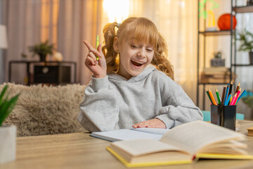 Young school girl sitting at table on sofa, focused on solving a task in notebook. Caucasian child...