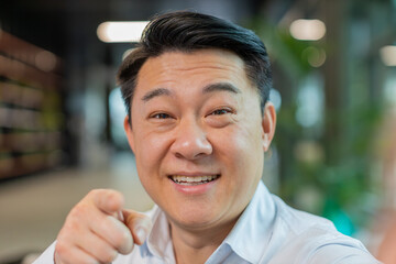 Fototapeta premium Closeup portrait of happy Chinese businessman boss shake hand hi gesture greeting welcomes someone, pointing to camera, online webinar, remote distance conversation. Entrepreneur man in office lobby.