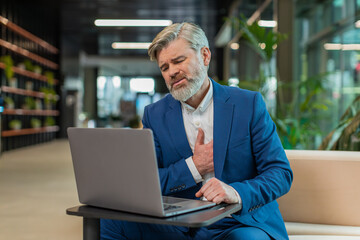 Stressed Caucasian mature businessman using laptop and suffers from tension holding chest with heart pain attack risk of stroke symptom while working in modern office lobby. Businessperson in suit.
