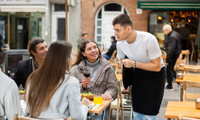 Open area of cafe, guy listen to restaurant couple of customer, write down order items in phone note, records order dish name in mobile application. Waiter of public catering answer customers question