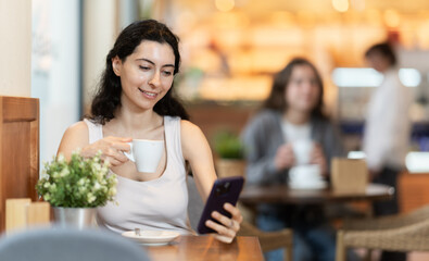 Young woman drinking hot black coffee and looking at phone screen, scrolling through social media feed and chatting with friends