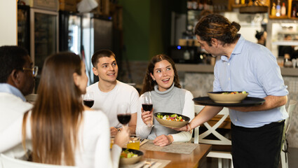 Male waiter delivers cooked food for a group of people in a European restaurant. Friends gathered in a restaurant and ordered delicious dishes. Customers receive quality service in the restaurant
