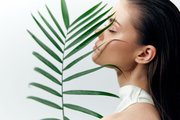 Naklejka premium Close-up portrait of a serene woman with long hair, gently touching a palm leaf, exuding calmness and nature-inspired beauty in a minimalist setting.