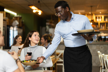 African waiter with a tray brings an order for a female client who is dining with a man. European woman having dinner in a restaurant