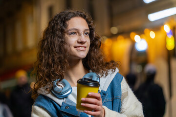 Happy tourist young woman enjoying coffee hot drink and smiling looking away thinking. Relaxing, taking a break. Teenager girl on urban city street, drinking coffee at night outdoors. Lifestyles.