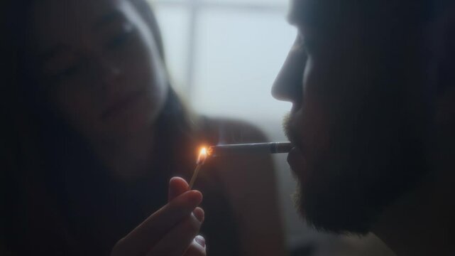 Side close-up shot of young woman with match lighting cigarette for her beloved spouse at home, affectionate love and understanding