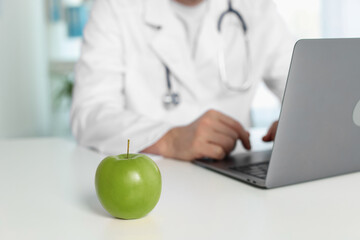 Nutritionist working with laptop at table in clinic, focus on green apple
