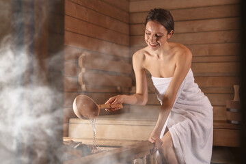 Smiling woman pouring water onto stones surrounded by steam in sauna