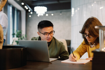 Asian man looking at female colleague drawing or writing in notebook