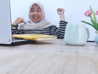 Asian woman wearing hijab with an enthusiastic expression celebrating an achievement with raising...