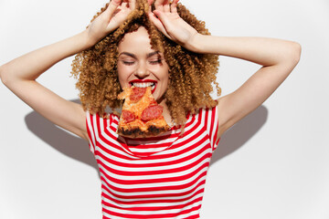 Happy young woman with curly hair enjoying pizza, wearing a striped shirt against a white...