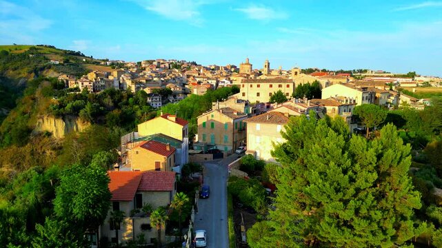 Peculiar aerial pedestal up shot from Offida, an ascending view with the town centre's buildings lying on a vegetation-filled hillscape, between roads, cars, roofs, bell towers, mostly clear azure sky