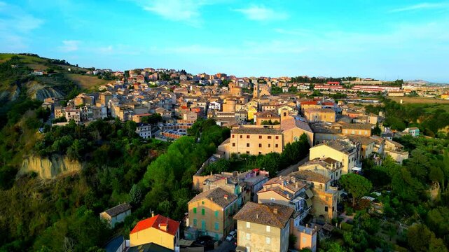 Calm aerial pedestal down shot from Offida, a descent amongst reddish roofs, vintage-styled buildings, roads, cars, bell towers, surrounding vegetation and the raw ground surface, slightly veiled sky