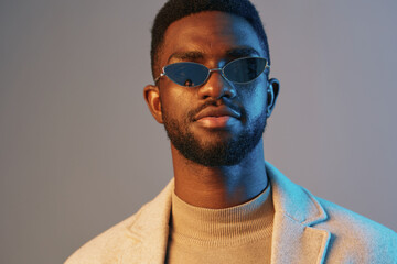 Stylish young African man wearing sunglasses and a beige turtleneck sweater, posing confidently against a two tone blue backdrop