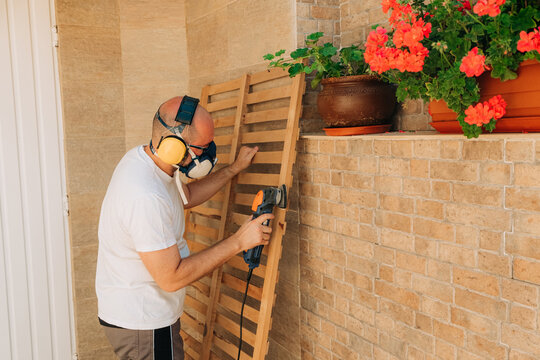 Man sanding wooden slats outdoors with protective gear - Powered by Adobe