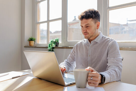 Focused office worker using laptop and drinking coffee at desk